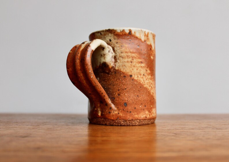 May include: A brown and white ceramic mug with a unique, twisted handle. The mug has a speckled glaze and a rustic look.