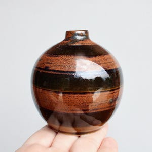 May include: A small, round ceramic vase with a narrow neck. The vase features horizontal bands of dark brown and reddish-brown glaze. The glossy finish reflects light, highlighting the color variations. The vase is held in a hand.