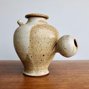 May include: A ceramic pitcher with a speckled brown and white glaze. The pitcher has a round body and a spout on the side. It also has a small, round handle on the side.