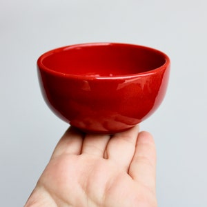 May include: A small, glossy red ceramic bowl. The bowl is round with a smooth, curved interior and exterior. The bowl is held in a person's hand against a plain white background. The bowl is a vibrant, solid red color.