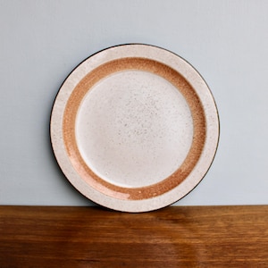 May include: A round ceramic plate with a speckled off-white center and a wide, rust-colored band around the edge. The plate has a dark brown rim and sits on a wooden surface. The plate is a piece of tableware.