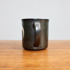 Pu&ograve; includere: Una tazza in ceramica verde scuro con un design bianco. La tazza ha un manico ed &egrave; appoggiata su una superficie di legno.