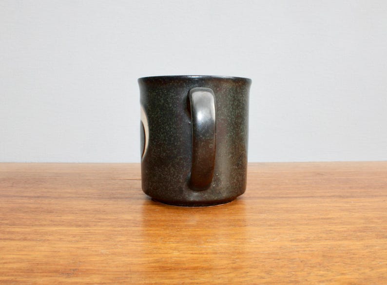 Pu&ograve; includere: Una tazza in ceramica verde scuro con un design bianco. La tazza ha un manico ed &egrave; appoggiata su una superficie di legno.