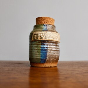 May include: A small, hand-crafted ceramic jar with a cork lid. The jar is decorated with a blue, green, and brown striped pattern and has the word "Grandma" written on the side.