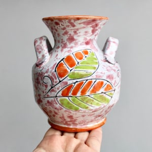 May include: A white ceramic vase with pink speckles and two handles. The vase is decorated with hand-painted orange and green leaves.