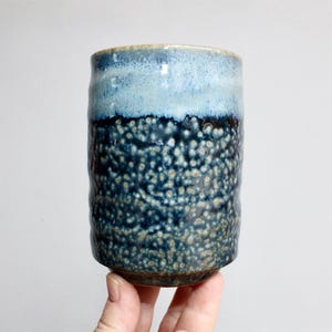 May include: A cylindrical ceramic tumbler with a textured surface. The top is light blue, transitioning to a dark blue speckled pattern. The handmade cup is held by a person's hand.