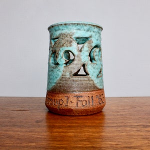 May include: A teal blue ceramic vase with cut-out designs and a brown base. The vase has the words "Group I - Fall '83" inscribed on the base.