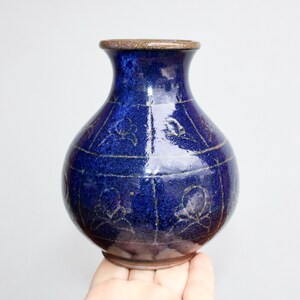 May include: A handcrafted, dark blue ceramic vase with a rounded body and a narrow neck. The vase features a textured surface with a pattern of lines and floral motifs. The rim is a contrasting brown color.