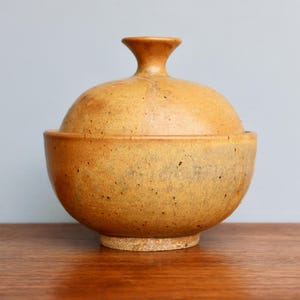 May include: A ceramic bowl with a lid, both in a speckled brown and yellow glaze. The bowl is round and has a slightly flared rim. The lid has a small knob on top.