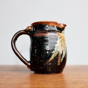 May include: A handmade ceramic pitcher with a dark brown and black speckled glaze. The pitcher has a rounded body, a curved handle, and a spout. The glaze features streaks of tan, green, and brown.