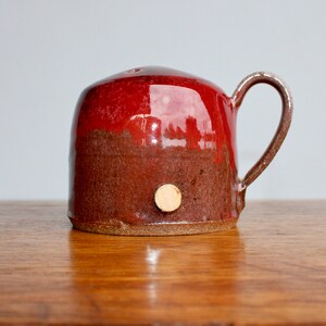 May include: A red and brown ceramic mug with a wooden knob on the side. The mug has a rounded shape and a handle.