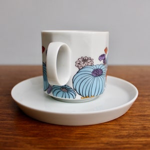 Może przedstawiać: Biały ceramiczny kubek z kwiatowym wzorem w kolorze niebieskim, fioletowym i pomarańczowym. Kubek stoi na pasującym białym spodku. Kubek ma zakrzywiony uchwyt. Zestaw stoi na drewnianej powierzchni.