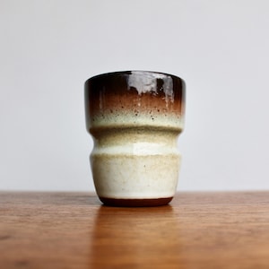 May include: A brown, white, and cream ceramic cup with a flared base. The cup has a glazed finish and a smooth, rounded shape.