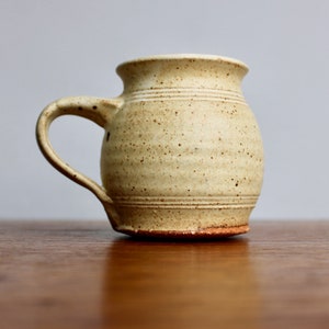 May include: A handmade ceramic mug with a speckled glaze in a light beige color. The mug has a rounded body and a handle.