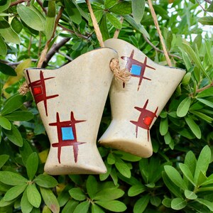 May include: Two cream-colored, vase-shaped ceramic planters with geometric red and blue designs, suspended by twine, hang against a backdrop of green foliage. The planters have a rustic, handmade appearance, adding a touch of bohemian charm.