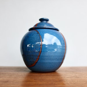 May include: A blue ceramic jar with a lid. The jar has a round shape with a glossy finish and a brown stripe design. The jar is sitting on a wooden surface.