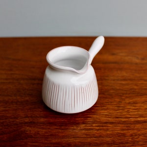 May include: White ceramic creamer with a small handle and a ribbed design. The creamer is on a wooden surface.