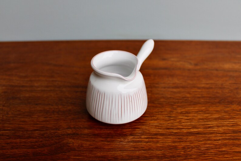 May include: White ceramic creamer with a small handle and a ribbed design. The creamer is on a wooden surface.