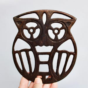 May include: A brown metal owl-shaped trivet with a cutout design. The trivet has a circular shape and is designed to protect surfaces from heat.