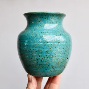 May include: A turquoise ceramic vase with a speckled glaze. The vase has a rounded body and a slightly flared opening.