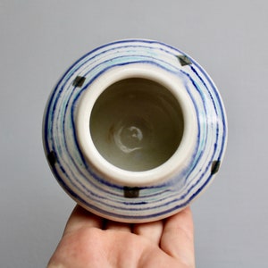 May include: A small white ceramic pot with blue and green stripes and black accents. The pot is hand-painted and has a round shape.