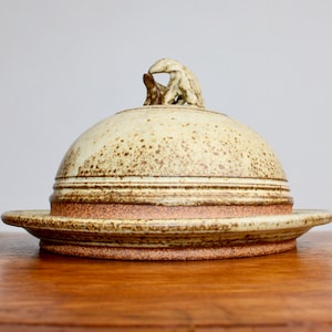 May include: A ceramic butter dish with a dome lid and a small animal figurine on top. The dish is glazed in a light brown color with brown speckles and has a brown rim.