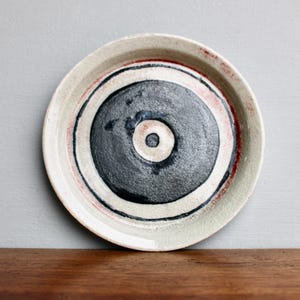 May include: A handmade ceramic plate with a circular design. The plate features concentric rings of black, white, and red against a cream-colored background. The center is a dark gray with a small white circle.