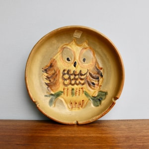 May include: A ceramic ashtray with a raised owl design. The owl is painted in shades of brown, yellow, and black. The ashtray is a light yellow color.
