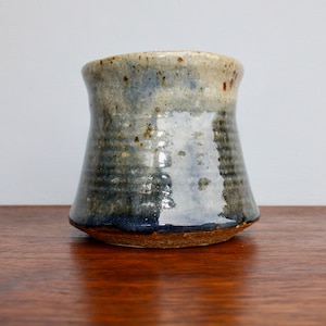 May include: A ceramic mug with a flared rim and a tapered base, featuring a glossy glaze that transitions from a speckled beige at the top to a deep, dark blue at the bottom, with visible horizontal ridges along the lower body of the vessel.