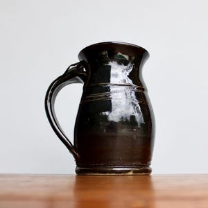 May include: A black ceramic mug with a rounded body and a handle. The mug has a glossy finish and a slight sheen.