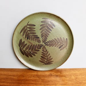 May include: A round ceramic plate with a green glaze and a brown fern design. The fern design is in a circular pattern.