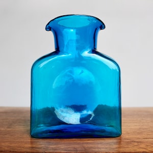 May include: A square-shaped, blue glass pitcher with a wide mouth and a small spout. The pitcher is made of clear glass with a slight blue tint.