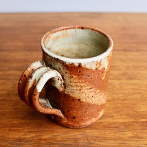 May include: A brown and white ceramic mug with a unique, sculpted handle. The mug has a speckled glaze and a rustic, handmade look.