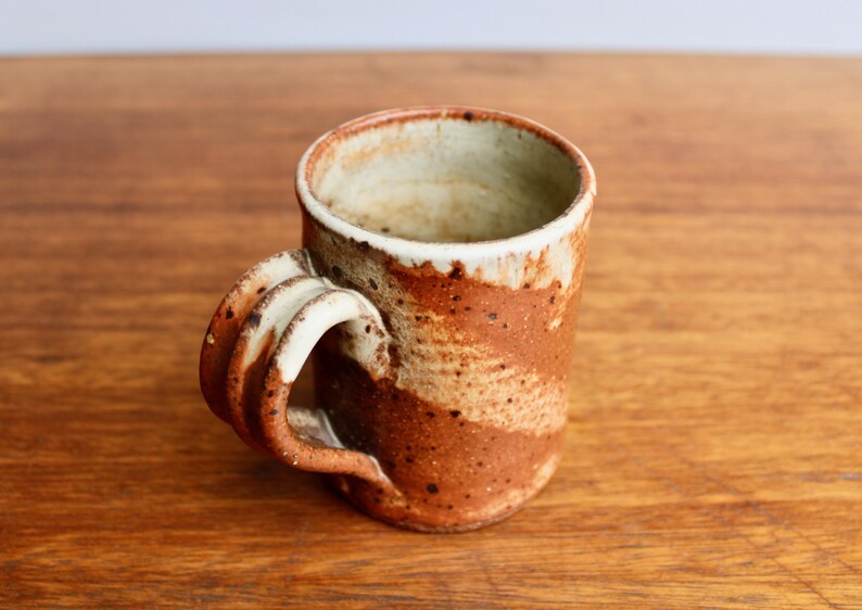 May include: A brown and white ceramic mug with a unique, sculpted handle. The mug has a speckled glaze and a rustic, handmade look.
