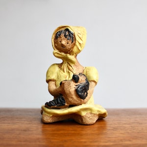 May include: A ceramic figurine of a person in a yellow dress and head covering, holding a brown object with black details. The figurine has a rustic, handmade appearance and sits on a wooden surface.