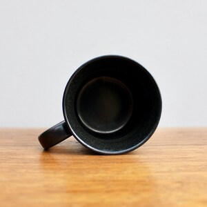 Pu&ograve; includere: Una tazza in ceramica nera con una base larga e arrotondata e un manico. La tazza &egrave; vuota e si trova su una superficie di legno.