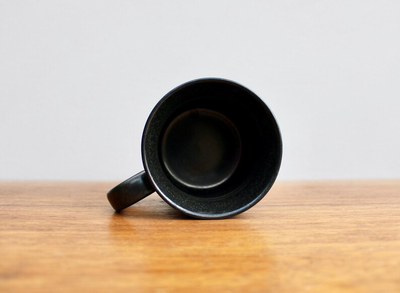 Pu&ograve; includere: Una tazza in ceramica nera con una base larga e arrotondata e un manico. La tazza &egrave; vuota e si trova su una superficie di legno.
