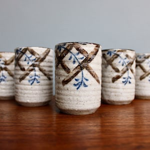 May include: Set of six white ceramic teacups with brown and blue floral designs. The cups have a textured surface and are arranged in a row on a wooden table.