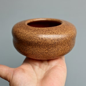 May include: A small, round, brown ceramic bowl with a speckled glaze. The bowl is sitting on a hand.