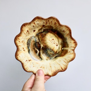 May include: A small, scalloped ceramic bowl with a white glaze and brown and yellow accents. The bowl has a unique, organic shape and a rustic, handmade feel.