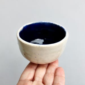 May include: A small white ceramic bowl with a dark blue interior. The bowl is slightly rounded and has a slightly uneven rim.