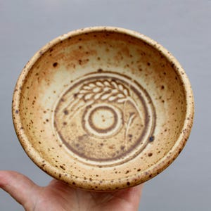 May include: A ceramic bowl with a speckled glaze in shades of brown and cream. The bowl has a raised design of wheat stalks in the center.