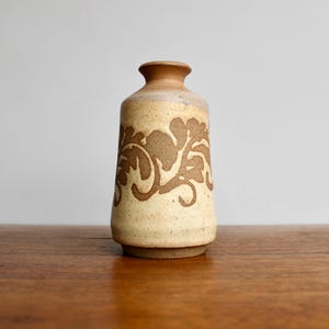 May include: A small, cream-colored ceramic vase with a brown floral design. The vase has a rounded base and a slightly flared opening.