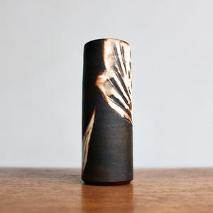 May include: A cylindrical ceramic vase with a dark brown exterior and a decorative white and brown pattern. The vase has a smooth, matte finish and stands on a wooden surface. The design is reminiscent of a stylized leaf.