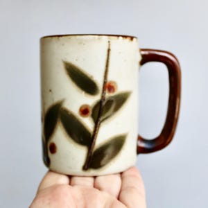 May include: A white ceramic mug with a brown handle and a leafy design in shades of brown and green. The leaves have small red berries.