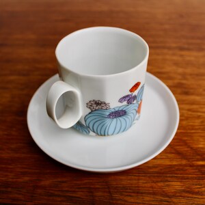Może przedstawiać: Biały ceramiczny zestaw filiżanki i spodka. Filiżanka ma kwiatowy wz&oacute;r z niebieskimi, fioletowymi i pomarańczowymi akcentami. Zestaw stoi na drewnianej powierzchni.