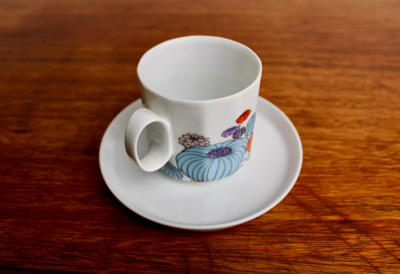 Może przedstawiać: Biały ceramiczny zestaw filiżanki i spodka. Filiżanka ma kwiatowy wz&oacute;r z niebieskimi, fioletowymi i pomarańczowymi akcentami. Zestaw stoi na drewnianej powierzchni.
