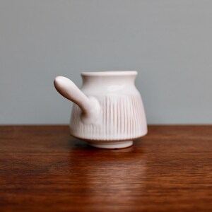 May include: White ceramic pitcher with a handle and vertical lines. The pitcher is on a wooden surface.