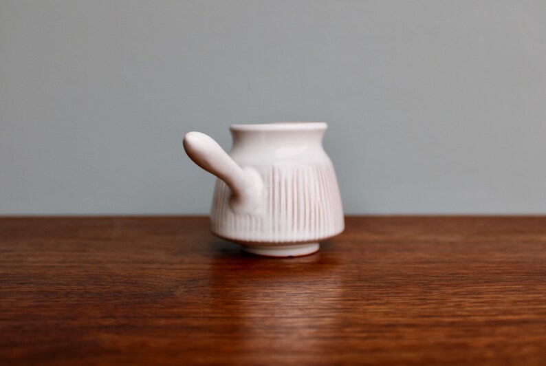 May include: White ceramic pitcher with a handle and vertical lines. The pitcher is on a wooden surface.