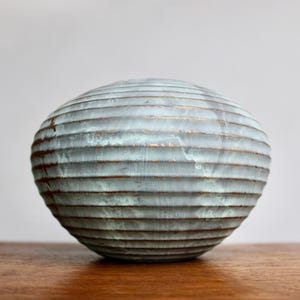 May include: A spherical, decorative ceramic vase with horizontal, textured lines. The vase has a light blue-green color with bronze-colored accents. The vase is sitting on a wooden surface.